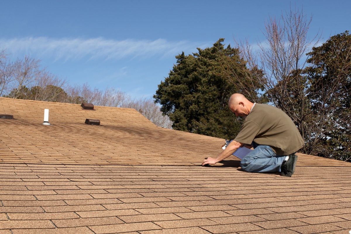 A man kneels on the roof of a house, focused on his task, with a clear sky in the background.