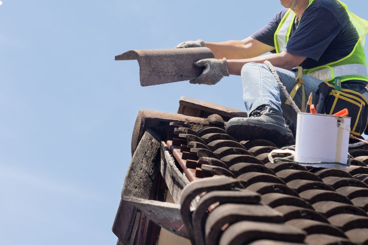 A man stands on a roof, holding a tool in one hand and a hammer in the other, ready for construction work.